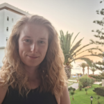 A blonde, white woman smiling at the camera at twilight. There is a white building and a palm tree behind her. She is wearing a dark strappy top and her hair is wavy.